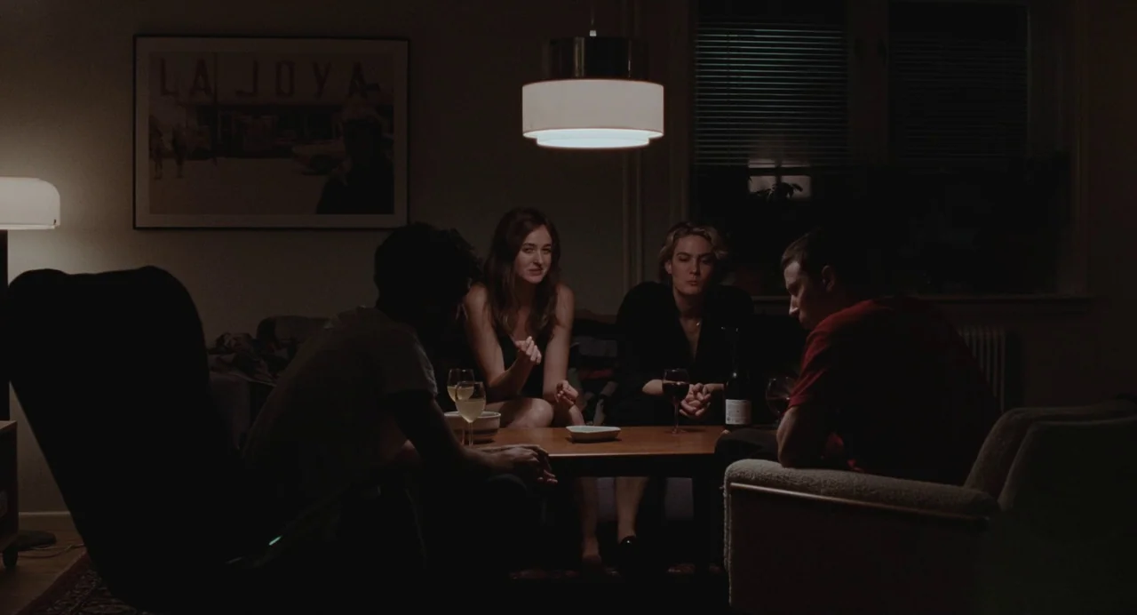 The Worst Person in the World (2021) movie still: wide — A dark living room features four people clustered around a low central wooden table. The…