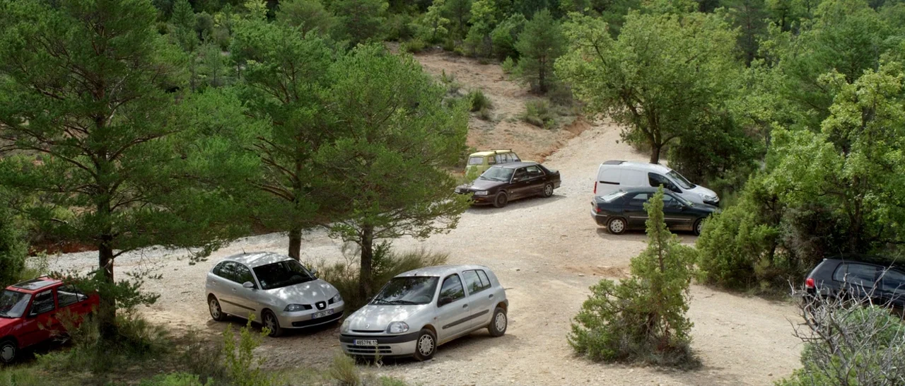 Stranger By The Lake (2013) movie still: wide — A gravel clearing situated within a dense forest of evergreen and deciduous trees. Several cars and…