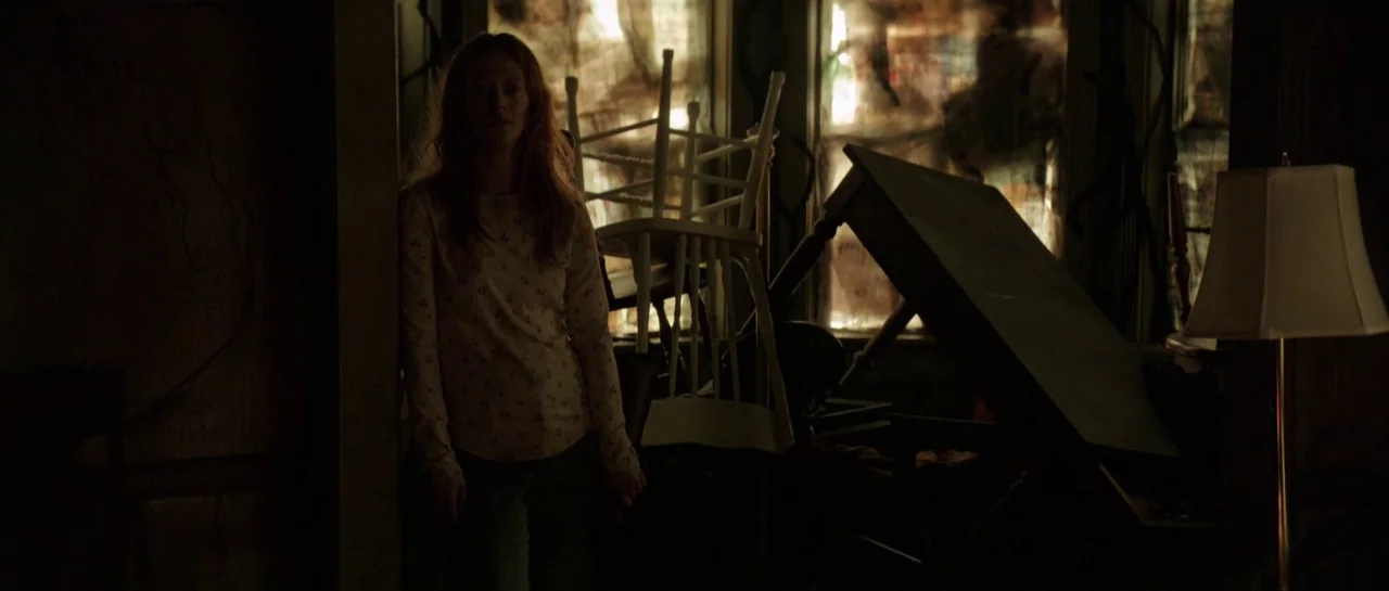 The Boogeyman (2023) movie still: medium — A dark, cluttered room with stacked wooden furniture and obscured windows. A woman stands in a narrow…