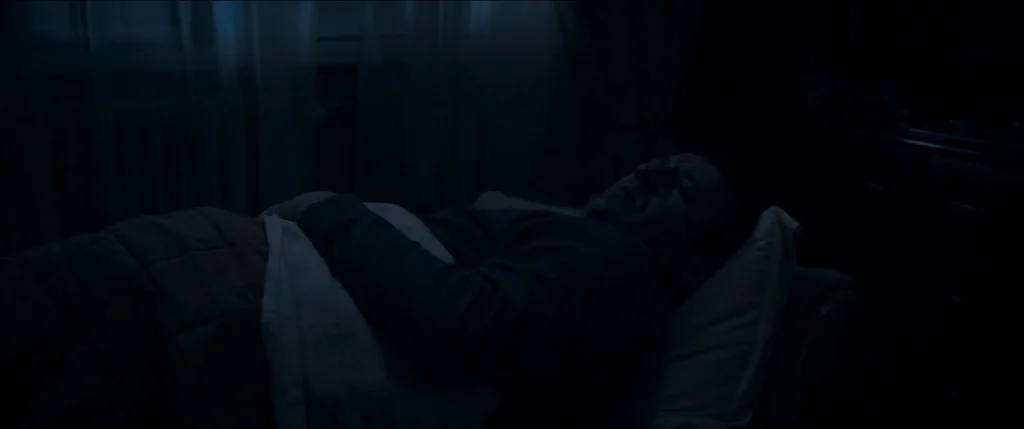 Graduation (2016) movie still: medium — A dark, dimly lit space with a person lying in bed near a window. The subject rests on a pillow covered by a…