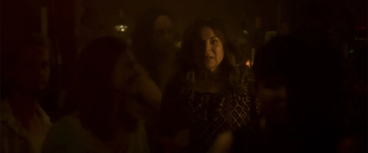 Jump, Darling (2022) movie still: medium — A dimly lit indoor gathering space with blurry silhouettes of people in the foreground. A woman stands in…