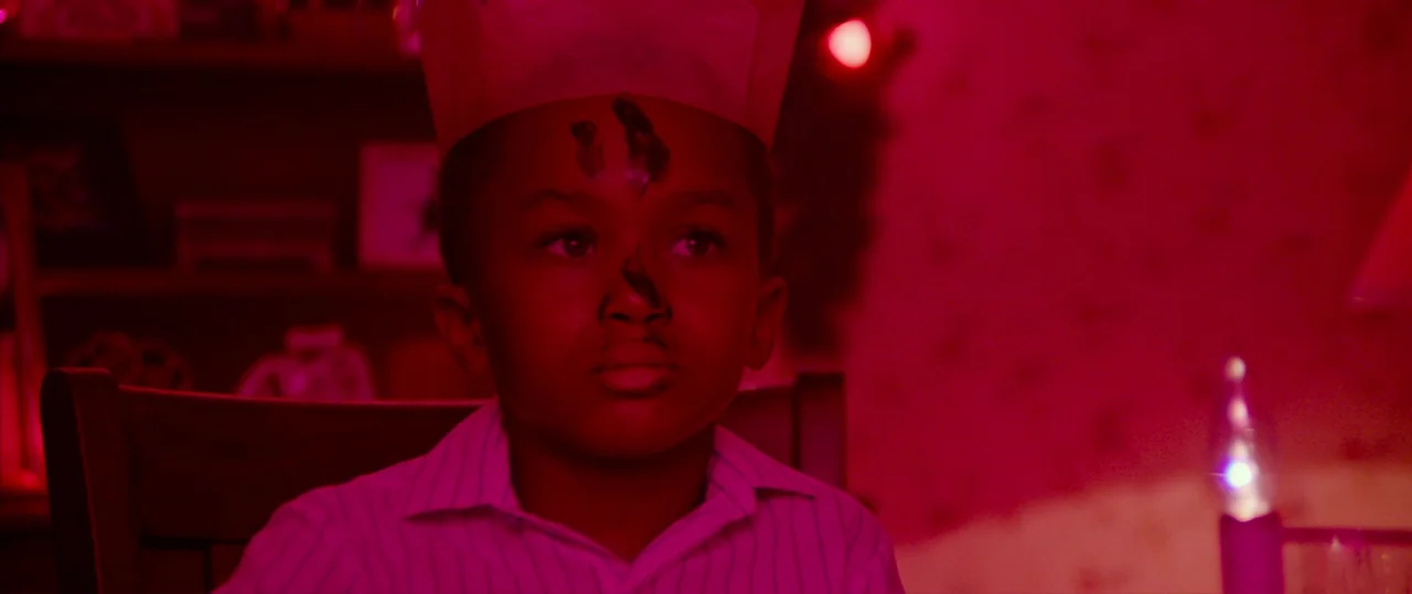 Random Acts of Violence (2019) movie still: closeup — A child wearing a paper crown sits at a table in a dimly lit room. He is bathed in a intense,…