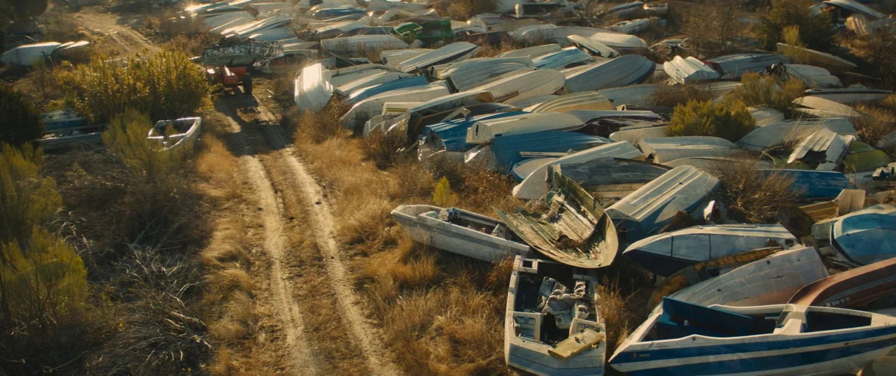 Manglehorn (2014) movie still: wide — A field filled with dozens of abandoned, weathered motorboats piled haphazardly in dry grass. A narrow, dusty…