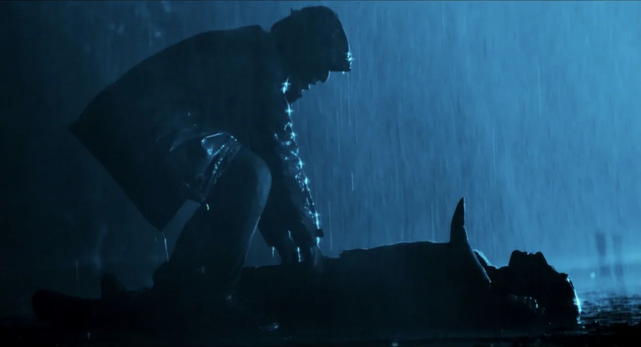 Echoes Of A Crime (2025) movie still: medium — A dark outdoor space at night with heavy rainfall falling on wet asphalt. One person kneels down over…