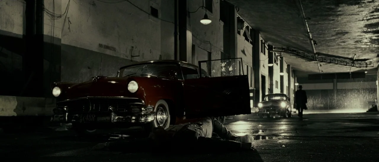 Slice (2018) movie still: wide — A dark, wet loading dock with a parked vintage red car and industrial walls. A man lies motionless on the ground as…