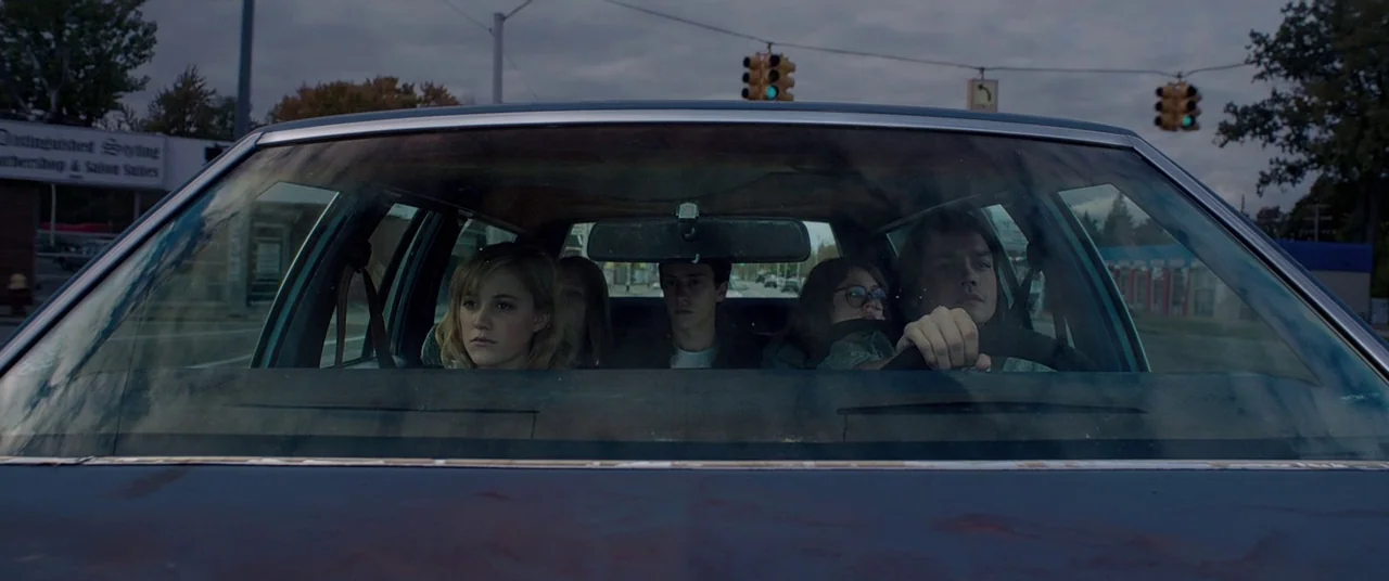 It Follows (2015) movie still: medium — Four young adults are inside a car driving on an overcast day. They sit together while looking ahead through…