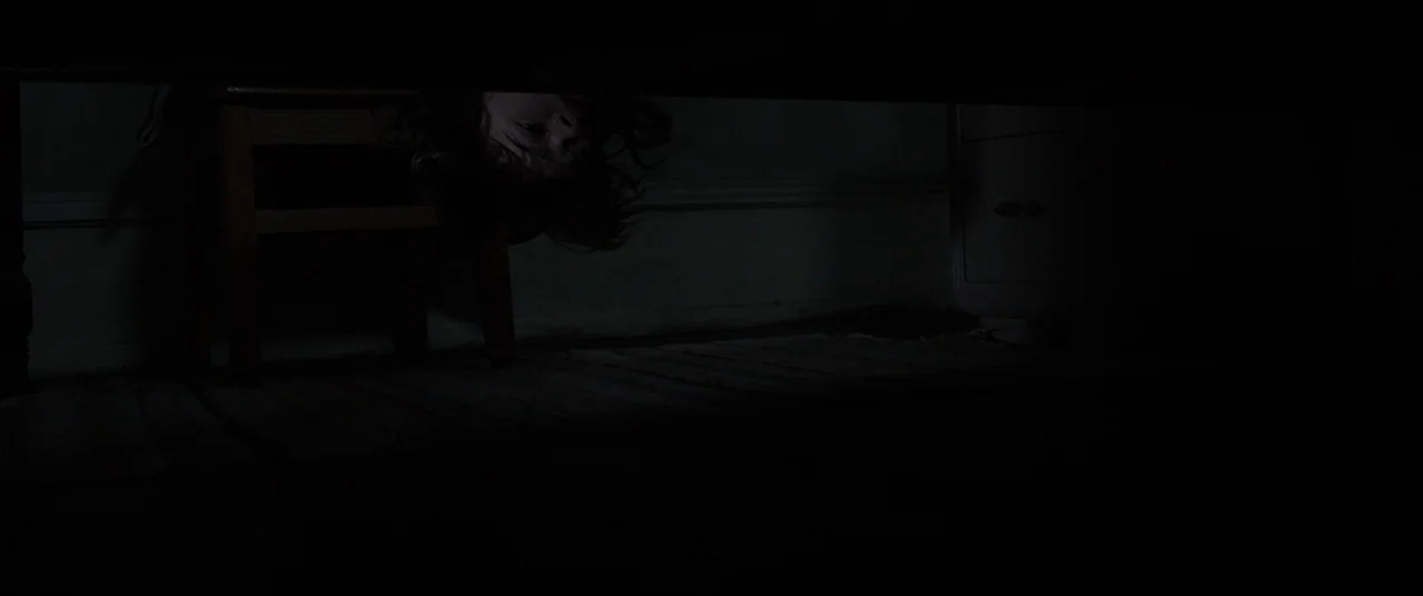 The Conjuring (2013) movie still: closeup — A dark, cramped space beneath a piece of wooden furniture in a room. A small child hangs their head down…