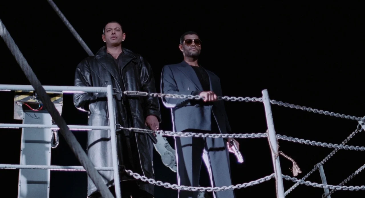 Deep Cover (1992) movie still: fullbody — Two men stand on a ship deck at night behind a metal chain railing. They hold pistols while looking…