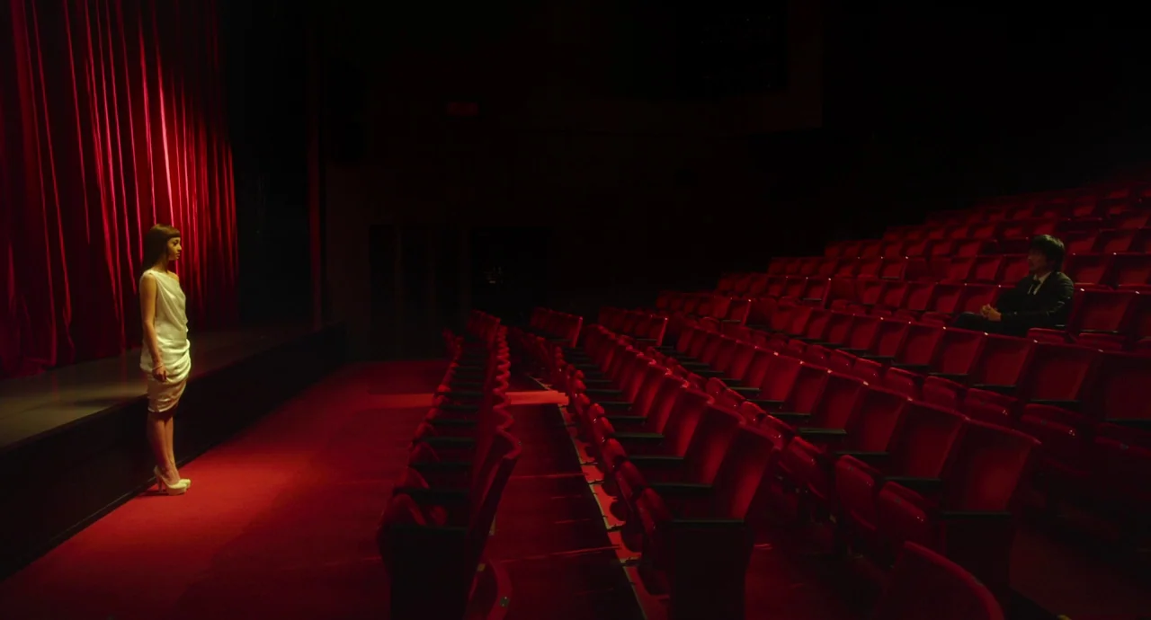 Helter Skelter (2012) movie still: wide — A dark, expansive auditorium features rows of empty red seats and a stage curtain. A woman stands alone on…
