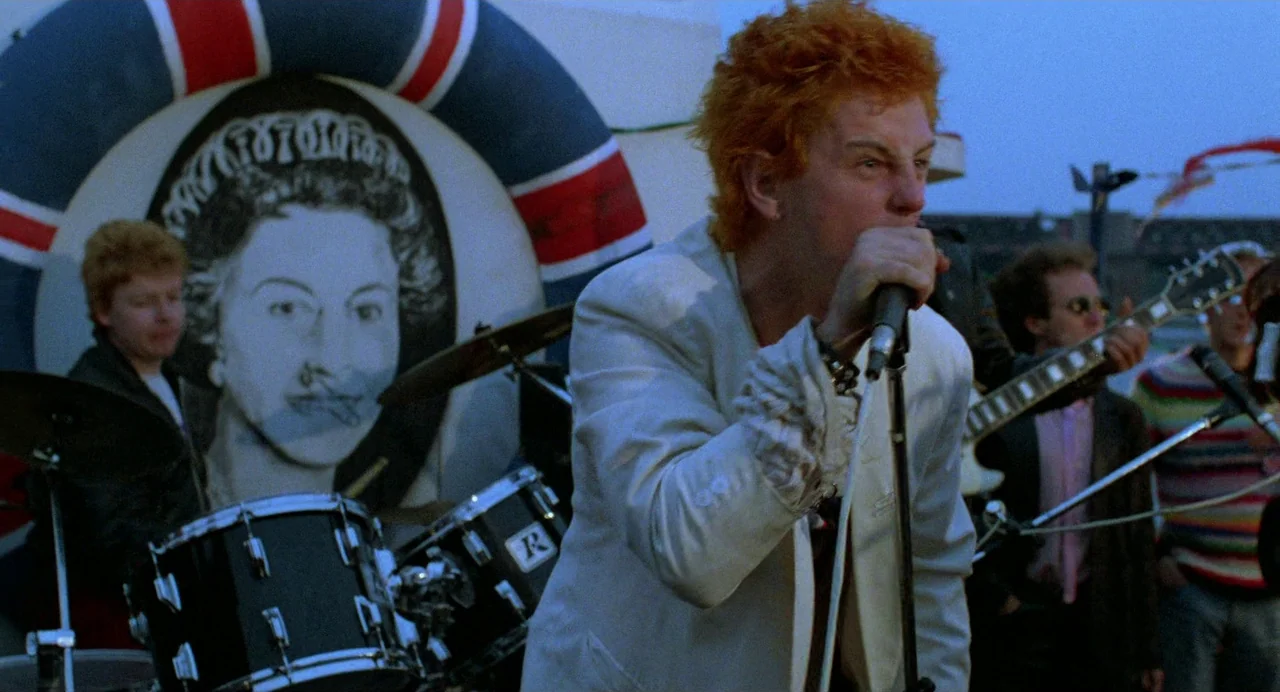 Sid and Nancy (1986) movie still: medium — A band performs outdoors on a flat roof or platform during an overcast day. A large circular banner…