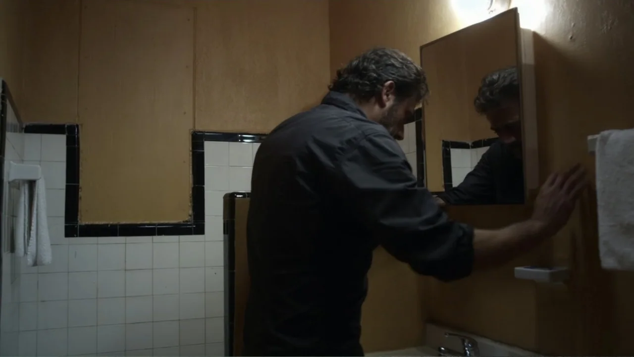 The Messenger (2015) movie still: medium — A small bathroom with beige walls and tiled wainscoting features a small medicine cabinet. A man stands…