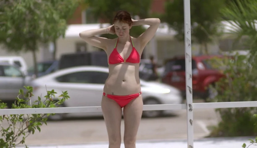 Kate Plays Christine (2016) movie still: fullbody — A woman stands in a sunny parking lot in front of a white railing. She holds both hands behind…
