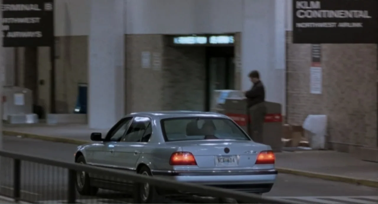 Forty Shades of Blue (2005) movie still: medium — An airport terminal drop-off area features beige brick pillars and informational signage. A man in…