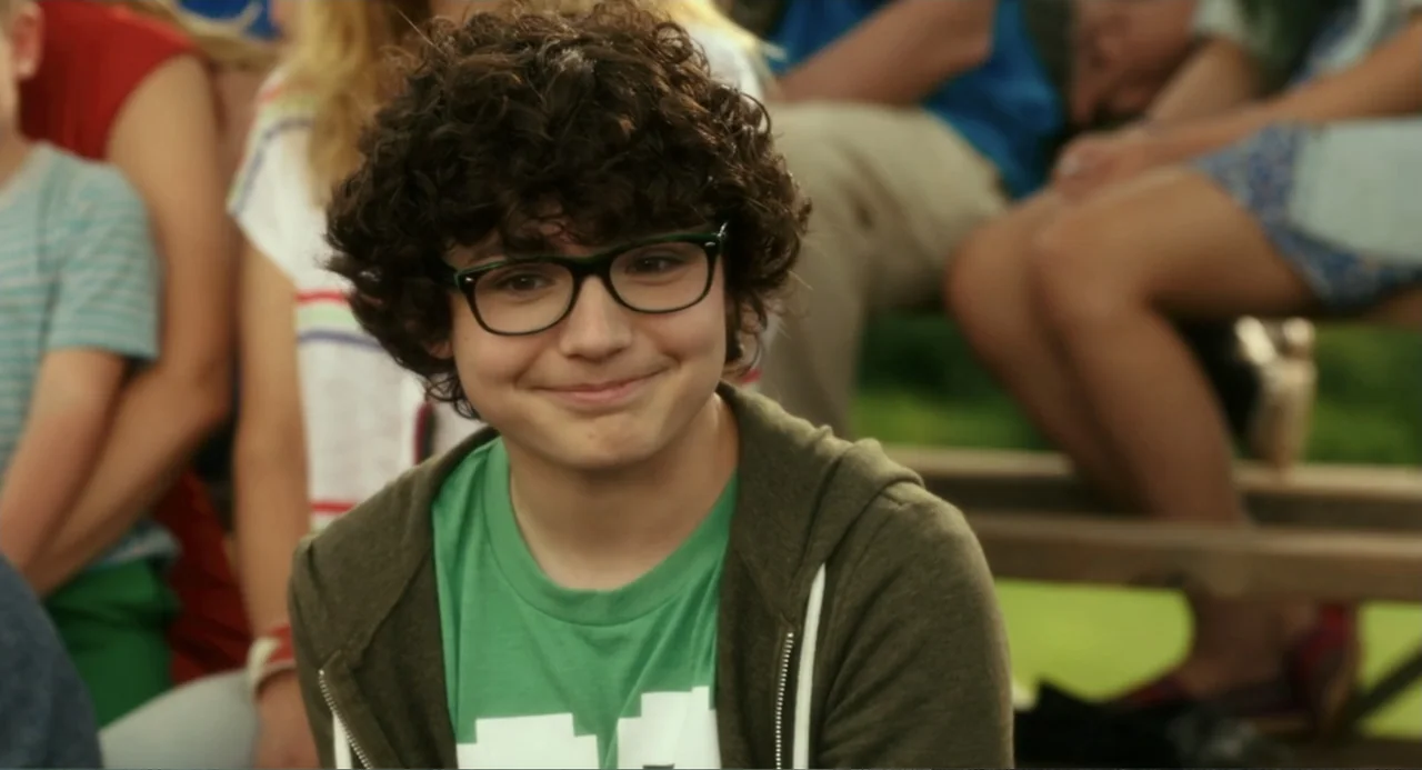 Blended (2014) movie still: medium — A teenage boy sits on wooden bleachers surrounded by blurred individuals in a park. He looks directly at the…