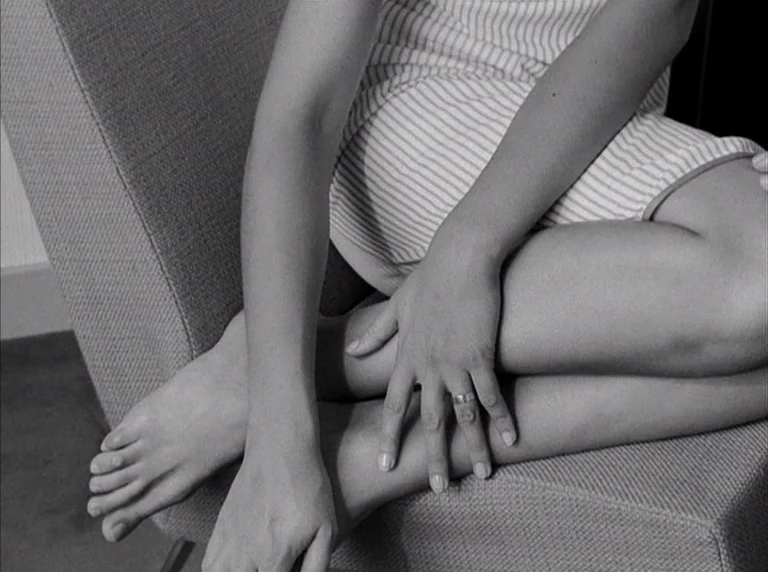 Une Femme Mariée (1964) movie still: closeup — A person sits on an upholstered piece of furniture with legs tucked together. The person's right hand…