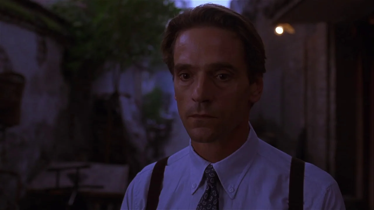 M. Butterfly (1993) movie still: medium — A man stands in a shadowed exterior courtyard during the late evening. He faces slightly away from the…