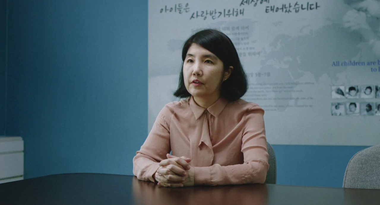 Return To Seoul (2022) movie still: medium — An office meeting room with blue walls and a large poster about children. A woman in a peach blouse…