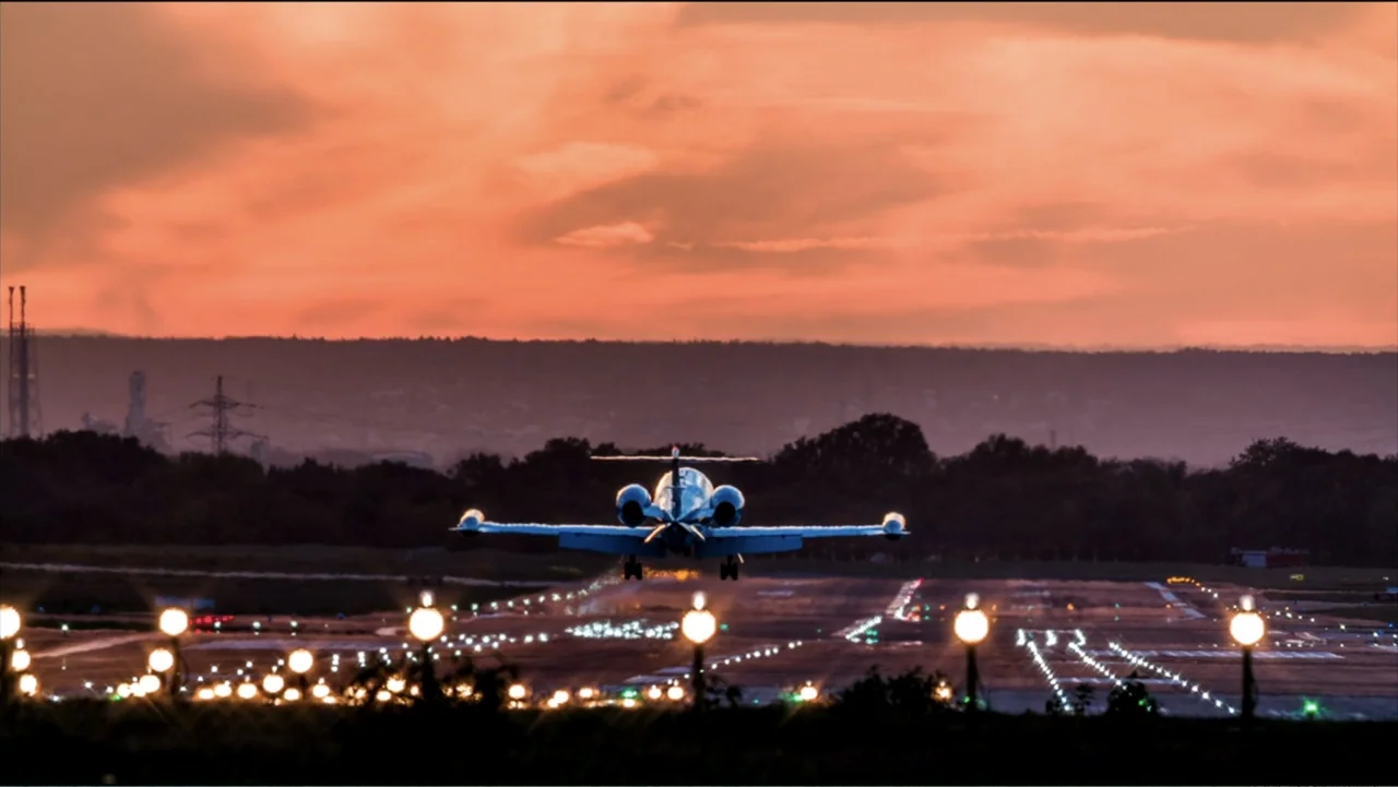 Jack in Time for Christmas (2024) movie still: wide — A private jet ascends from an airport runway against a vibrant sunset sky. The airfield is…