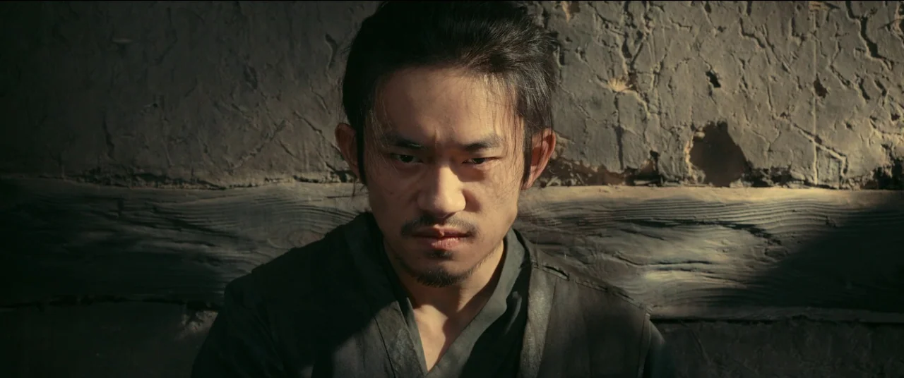 Full River Red (2023) movie still: closeup — A man stands before a weathered, textured wooden wall with visible cracks and grain. The man stares…