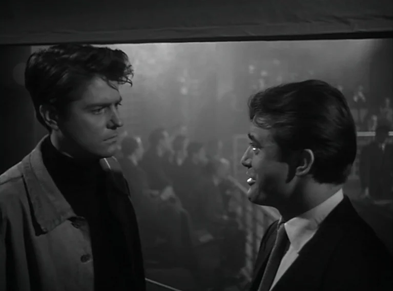 Head Against The Wall (1959) movie still: medium — Two men stand facing each other in a dimly lit, smoky room. A blurred audience is visible sitting…
