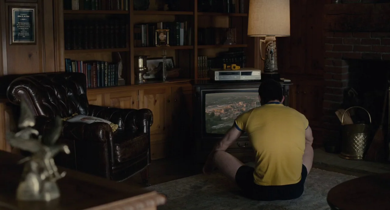 Foxcatcher (2014) movie still: medium — A wood-paneled room features a fireplace, a leather armchair, and many shelves of books. A man wearing a…