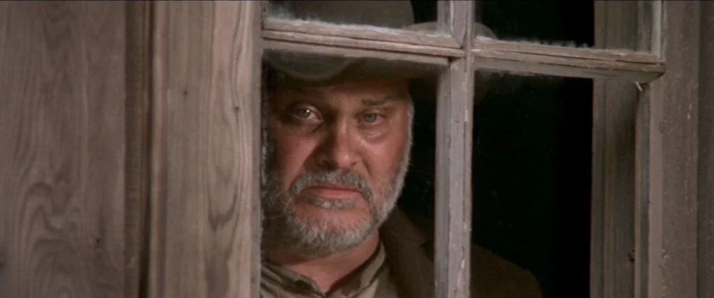In A Valley of Violence (2016) movie still: closeup — An older man peers through the divided panes of a wooden window frame. The man wears a brown…