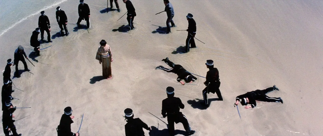 Lady Snowblood Song of Vengeance (1974) movie still: wide — A wide, high-angle shot depicts a group of men in black uniforms on sand. The men…