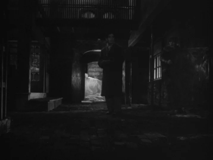 The Idiot (1951) movie still: fullbody — A dark urban alleyway features a central archway illuminated by a bright light source. A man in a long coat…