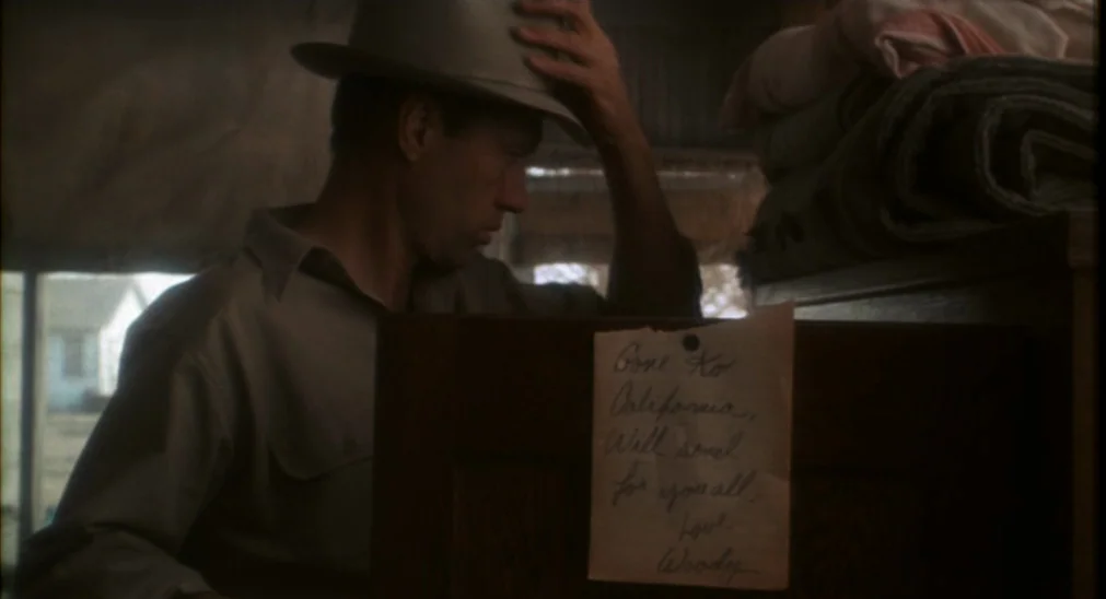 Bound For Glory (1976) movie still: medium — A dimly lit room with wooden furniture and a stack of folded blankets. A man stands wearing a fedora…