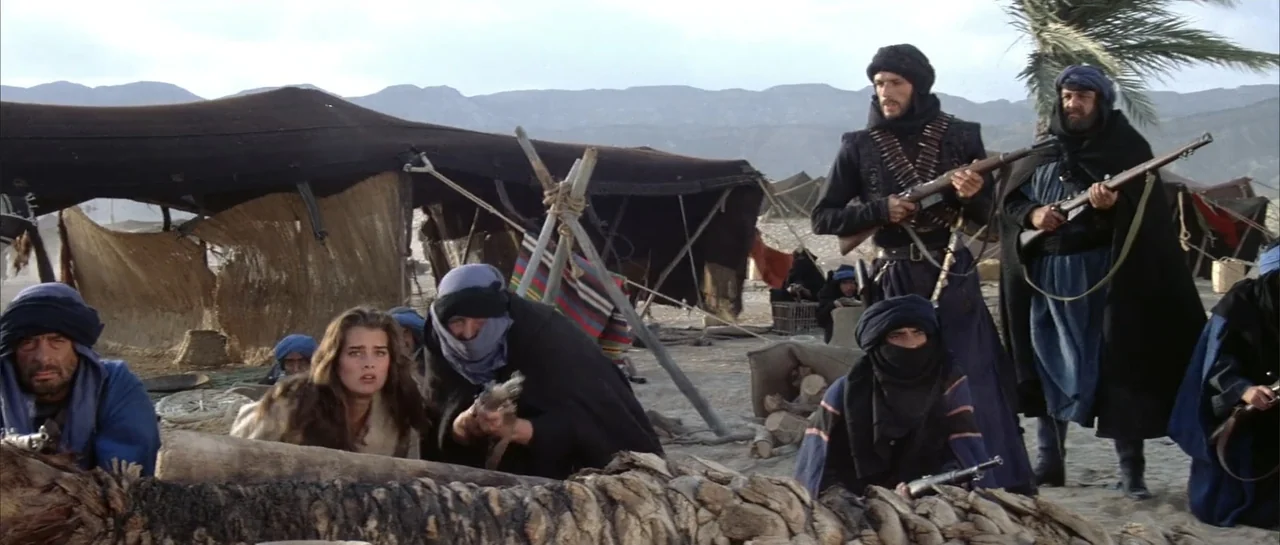 Sahara (1995) movie still: wide — A desert encampment features dark canvas tents and scattered equipment against a distant mountain range. A group…