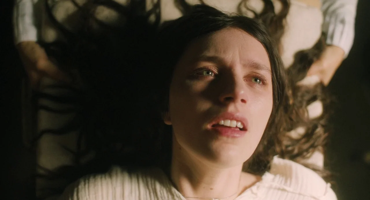 The First Omen (2024) movie still: closeup — A person lies on a pale surface with their dark hair fanned out. They look upward with their mouth…