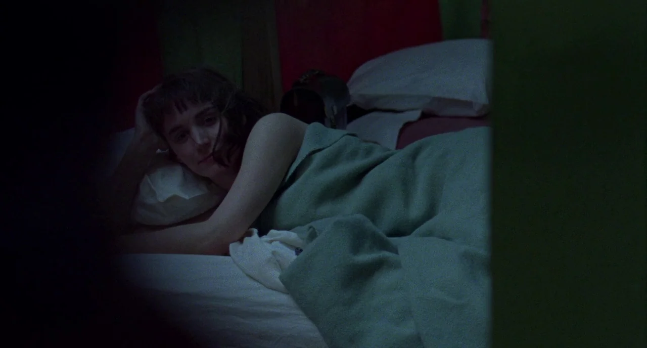Carol (2015) movie still: medium — A dark bedroom with red and green walls features a bed with white linens. A woman with short dark hair rests on…