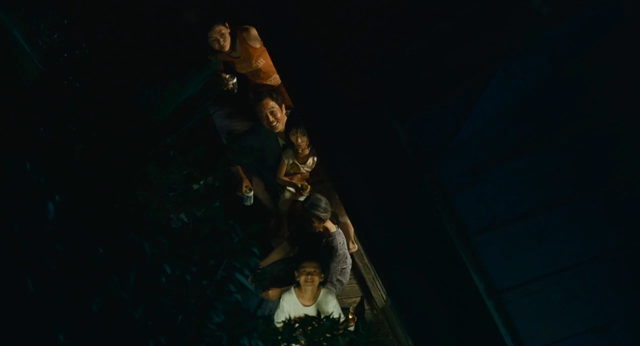 Shoplifters (2018) movie still: wide — A group of people sits on a wooden porch surrounded by dark vegetation. They look directly upward toward the…