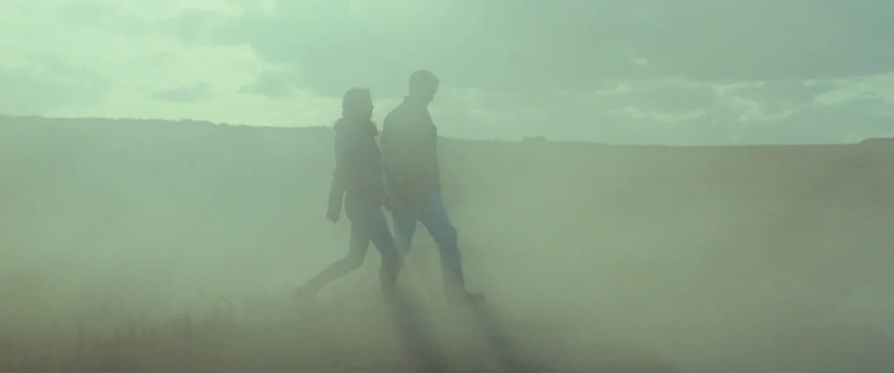 Catch Me Daddy (2014) movie still: wide — An open rural field is filled with thick airborne dust particles. A man and woman walk together from left…