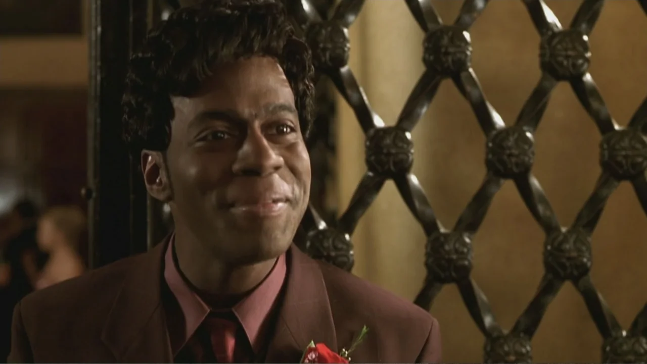 Not Another Teen Movie (2001) movie still: closeup — A man in a suit stands before a decorative metal gate in a hallway. He smiles as he faces the…