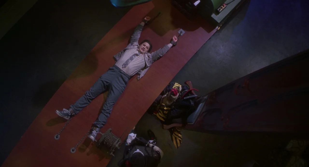 Monkeybone (2001) movie still: wide — A man is restrained by chains on a large wooden table inside a dimly lit space. An individual in a mask stands…
