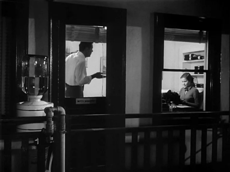 Sunset Boulevard (1950) movie still: wide — Two small glass-enclosed cubicles sit along a dark hallway on a studio soundstage. A man stands in the…