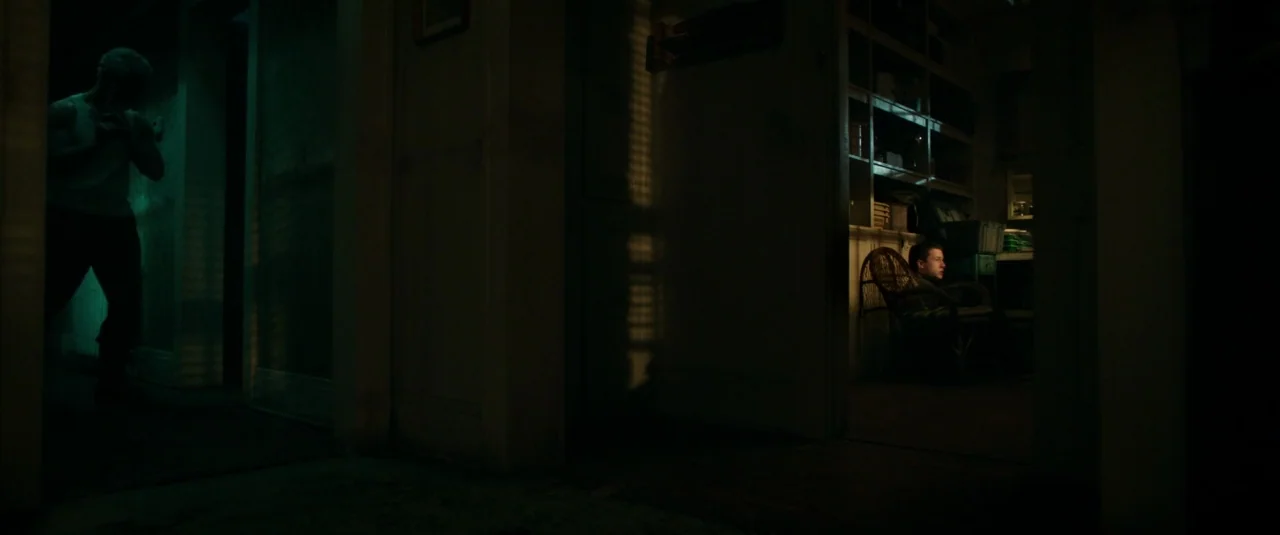 Don’t Breathe (2016) movie still: wide — A dark house interior with a long hallway connecting two rooms in low light. One man stands in the left…