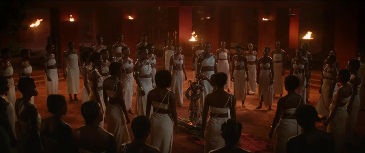 The Woman King (2022) movie still: wide — A large, open-air courtyard with red clay ground is illuminated by flickering torches. Many women wearing…