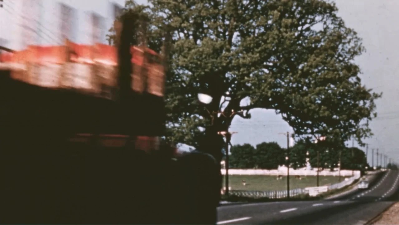Reggie (2023) movie still: wide — A rural paved road stretches into the distance beside a green meadow and trees. A moving vehicle is captured in…