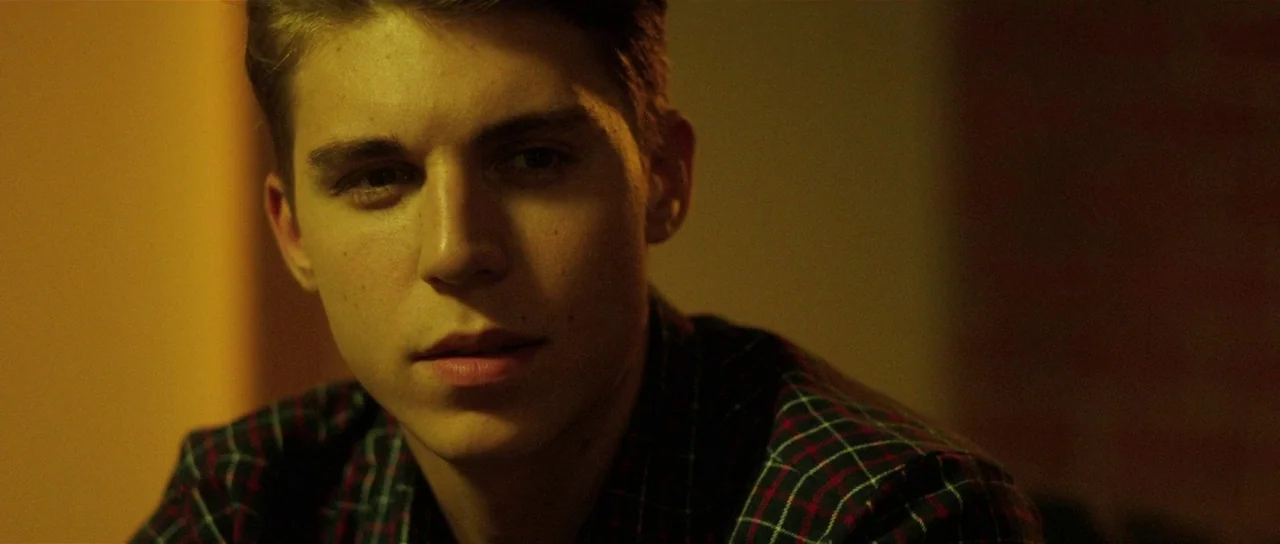 The Canyons (2013) movie still: closeup — A dimly lit room with warm yellow artificial lighting against a dark wall. A young man looks towards the…