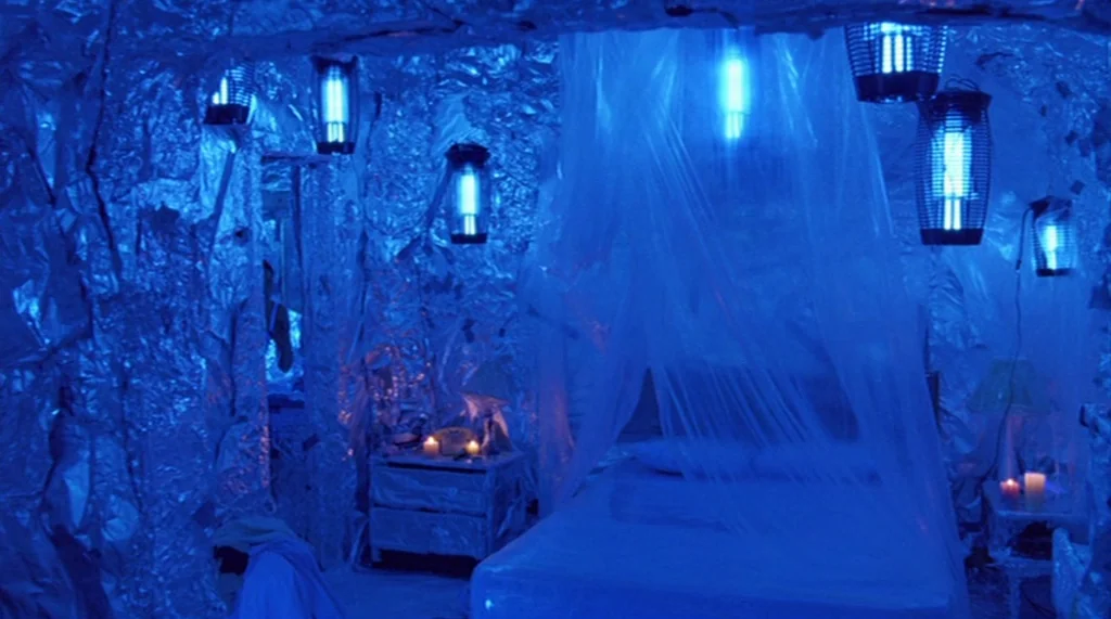 Bug movie still: wide — A small room has all walls and furniture wrapped in wrinkled silver aluminum foil. A bed sits in the center with a sheer…