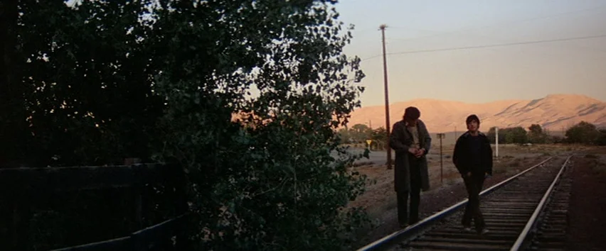 Scarecrow (1973) movie still: wide — A rural landscape with railroad tracks extending toward distant mountains at golden hour. Two men walk along…