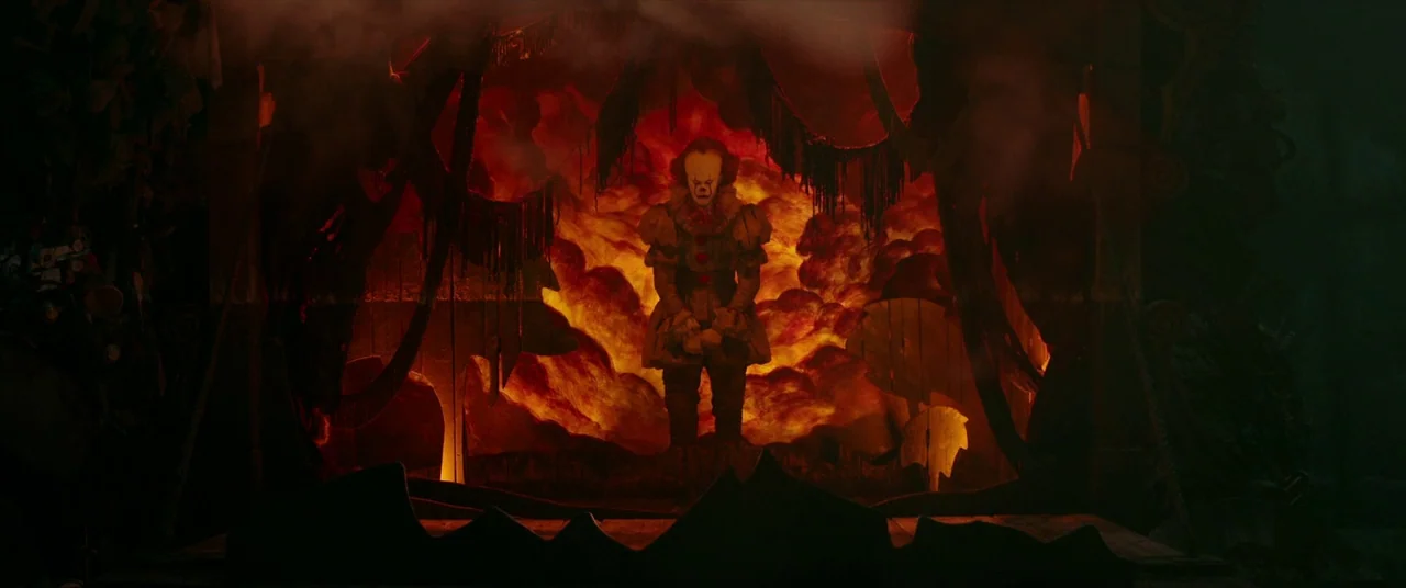 It (2017) movie still: fullbody — A dark wooden puppet booth is filled with heavy smoke and bright orange flames. A pale clown figure stands…