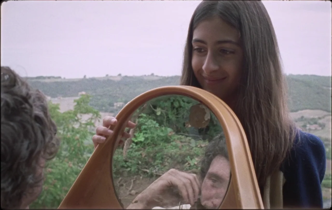 La Chimera (2023) movie still: medium — A young woman stands outdoors on a grassy hillside with a rolling landscape background. She holds a circular…