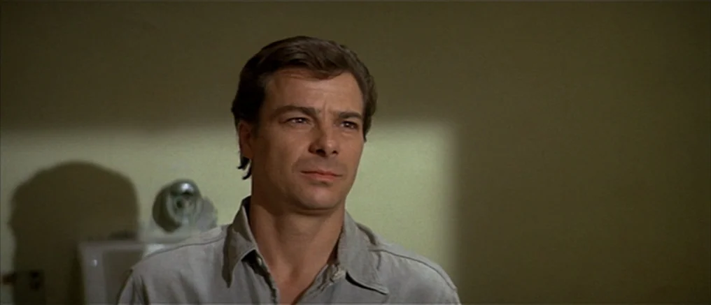 Assault on Precinct 13 (1976) movie still: closeup — An interior wall with a muted yellow hue forms the backdrop behind a man. The man stands facing…
