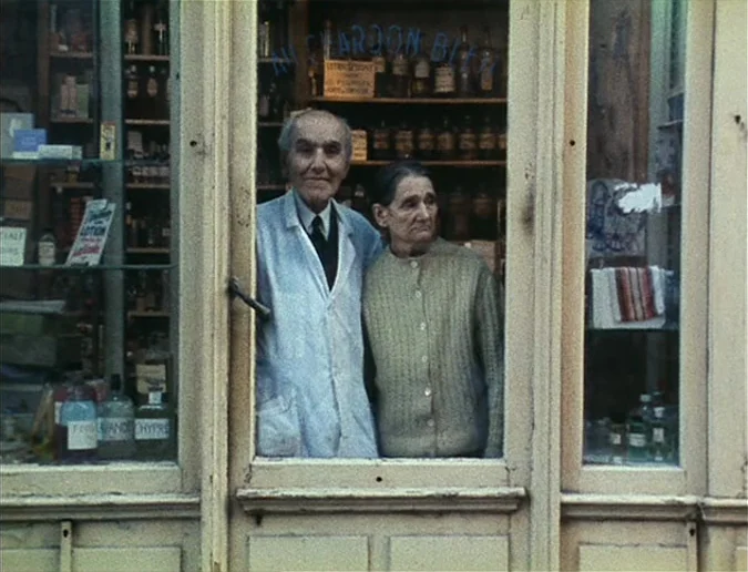 Daguerréotypes (1976) movie still: medium — An elderly couple stands behind the glass entrance of a vintage pharmacy storefront. Shelves filled with…