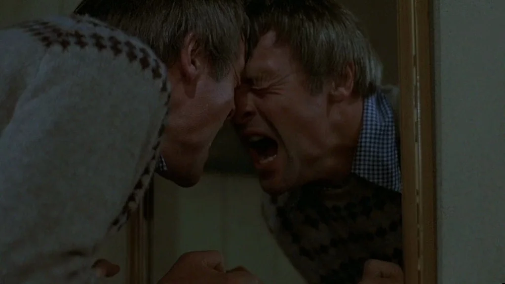 Magic (1978) movie still: closeup — A dark, cramped bathroom interior with a mirror reflecting a man's face. The man leans forward until his…