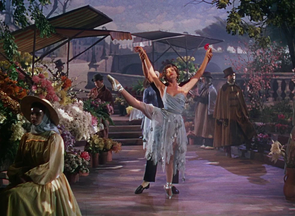 An American In Paris (1951) movie still: fullbody — An outdoor flower market set with painted backdrop of a bridge in Paris. A woman in a blue…