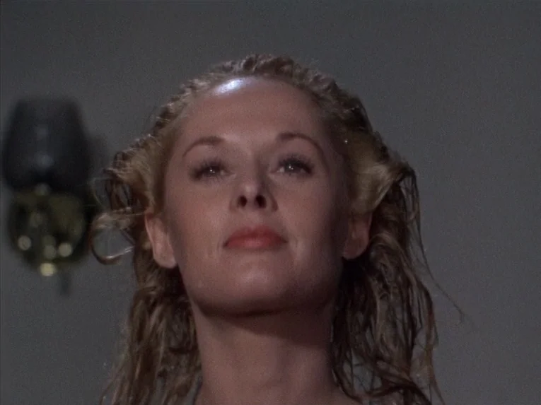 Marnie (1964) movie still: closeup — An interior bathroom with a blurred, dimly lit wall fixture in the background. A woman with wet hair looks…